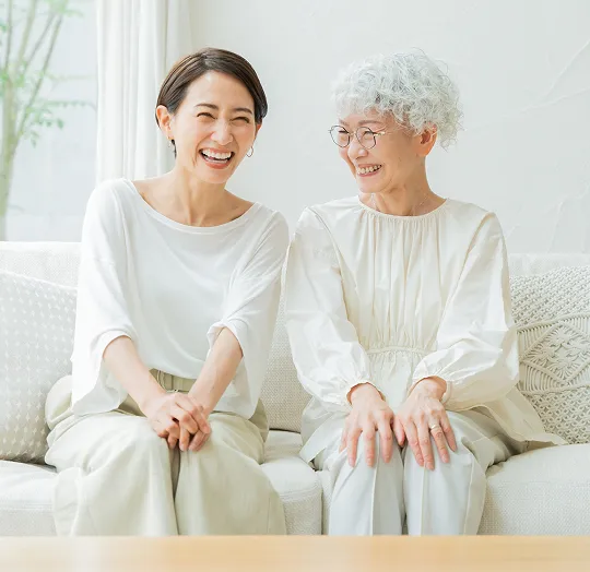 Two women, one younger and one older, sitting on a white couch smiling and engaging warmly.