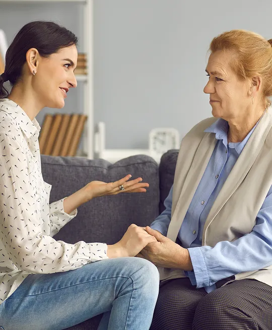 Two women seated on a gray sofa, engaged in a heartfelt conversation, holding hands.