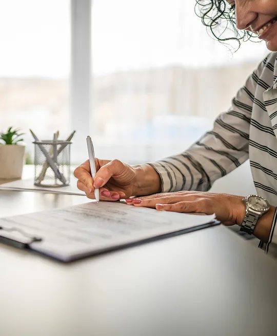 Person wearing a striped shirt writing on a clipboard with a pen at a desk.