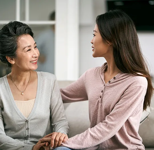 Two Asian women, one older and one younger, sitting and holding hands while smiling indoors.