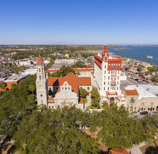 Aerial view of historic buildings with red roofs and a coastal cityscape under a clear blue sky.