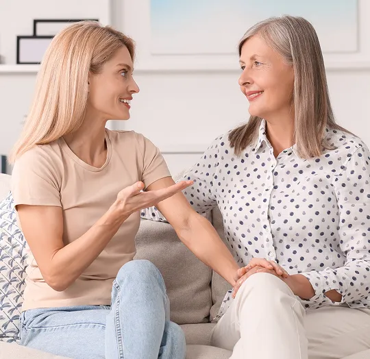 Two women sitting on a couch, engaged in a warm conversation, one holding the other's hand.