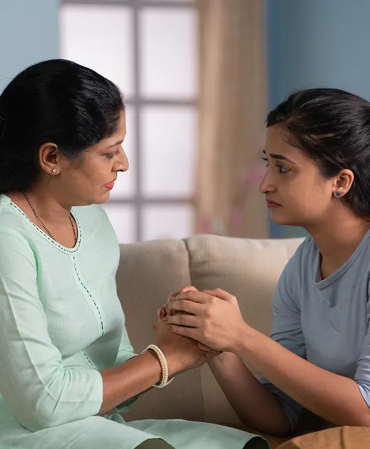 Two women sitting on a beige sofa, holding hands and engaging in a serious conversation.