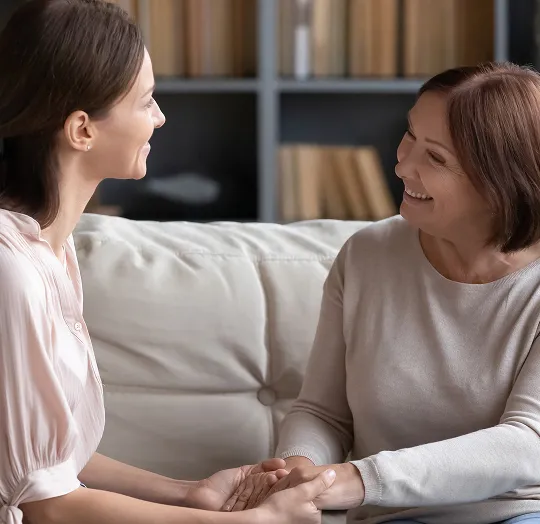 Two women sitting on a beige couch, smiling and holding hands in a warmly lit room.