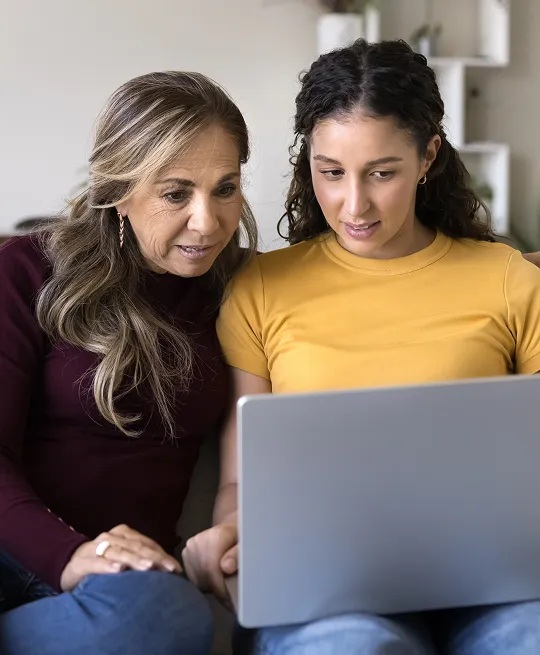 Two women, one in a maroon top and the other in a yellow shirt, looking at a laptop screen together.