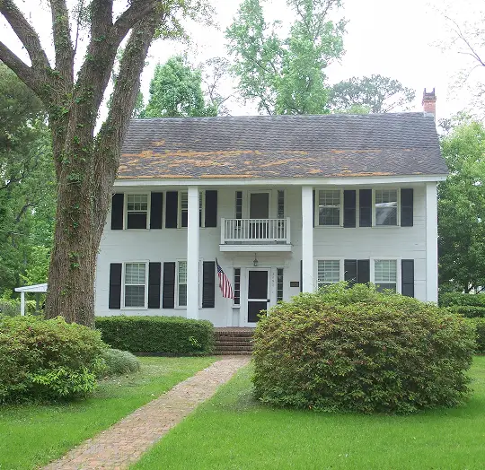 Two-story white colonial-style house with black shutters, a central balcony, American flag, surrounded by green trees and shrubs.