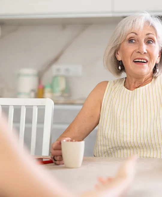 Elderly woman with gray hair wearing a striped sleeveless top, sitting at a table with a white mug in a modern kitchen.