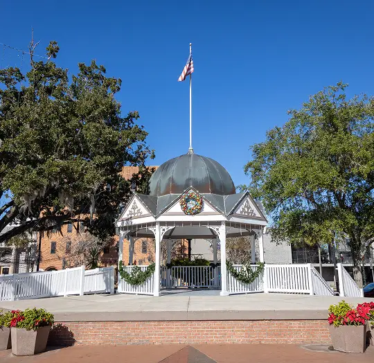 White gazebo with a domed roof, American flag, and holiday wreaths, surrounded by trees.