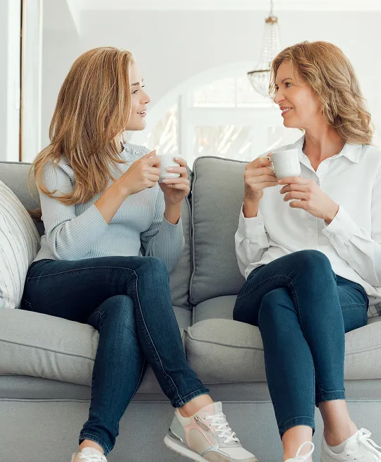 Two women seated on a gray sofa, holding white mugs and engaged in conversation.