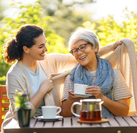 Two women, one younger and one older, share a blanket and smile while drinking tea outdoors.