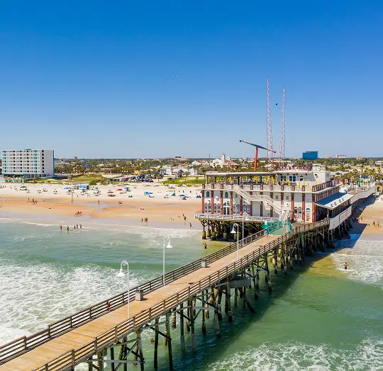 A wooden pier extending over the ocean with a large building at the end, a sandy beach with people and umbrellas, and a clear blue sky.