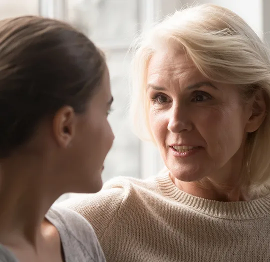 Two women engaged in a close, attentive conversation near a window with natural light.