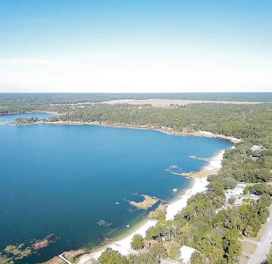 Aerial view of a large blue lake bordered by a sandy shoreline and dense green forest under a clear sky.
