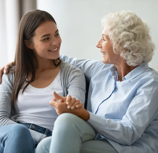 Two women, one elderly with white curly hair and one younger with long brown hair, smiling and holding hands while sitting on a couch.