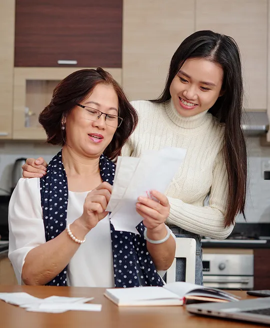 Two women in a kitchen reviewing documents, with one seated and the other standing behind her.