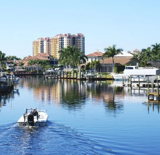 Small motorboat navigating a calm waterway lined with palm trees, residential houses, and a tall condominium building.