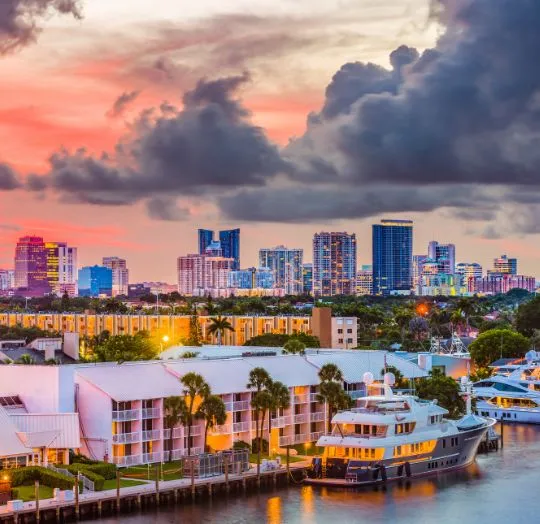 City skyline at sunset with high-rise buildings, waterfront residences, palm trees, and yachts docked in marina.