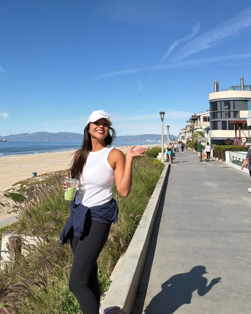 Woman in white tank top and black leggings holding a green drink, standing on a beachside promenade.