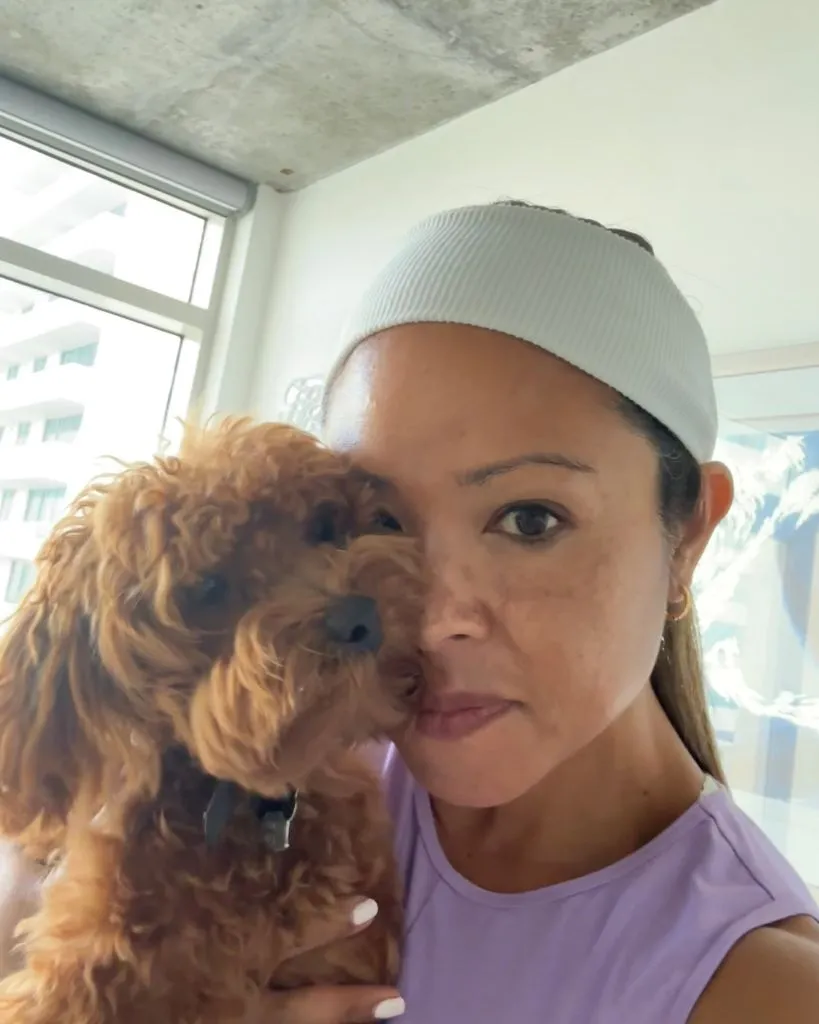 Woman with light purple sleeveless top and white headband holding a small brown curly-haired dog indoors.
