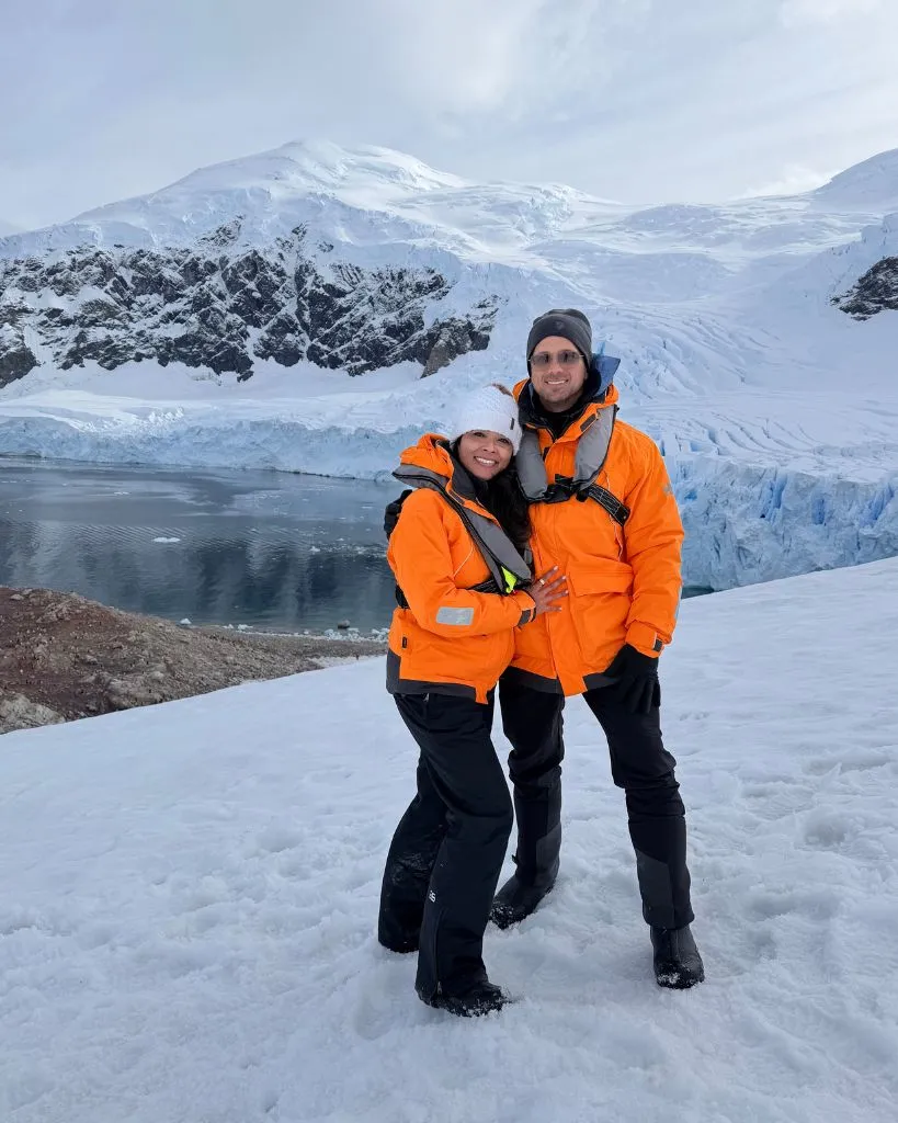Two people wearing bright orange jackets and winter gear standing on snow near a frozen lake with snowy mountains in the background.