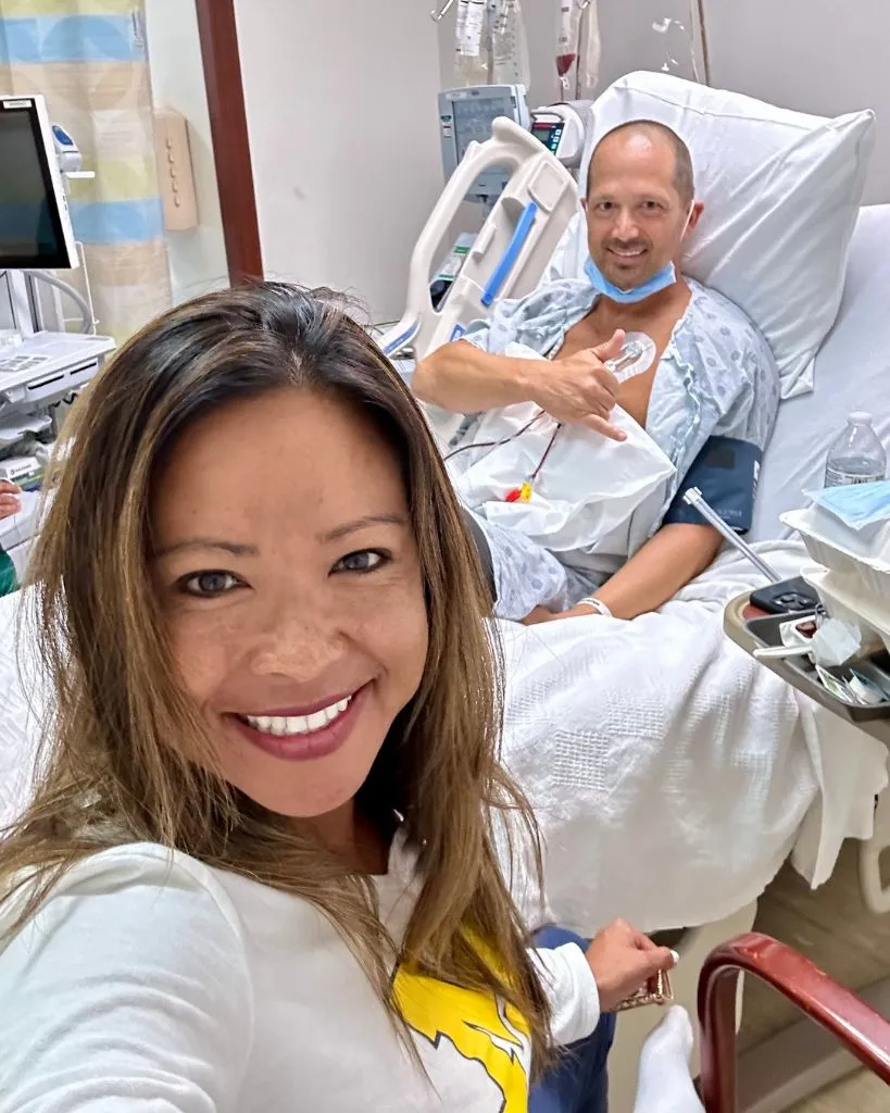 Smiling woman taking selfie with man in hospital bed giving hand gesture.