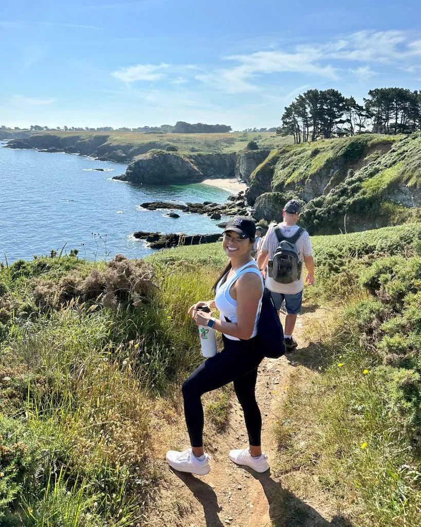 Two hikers on a coastal trail with cliffs, greenery, and a blue ocean under a clear sky.