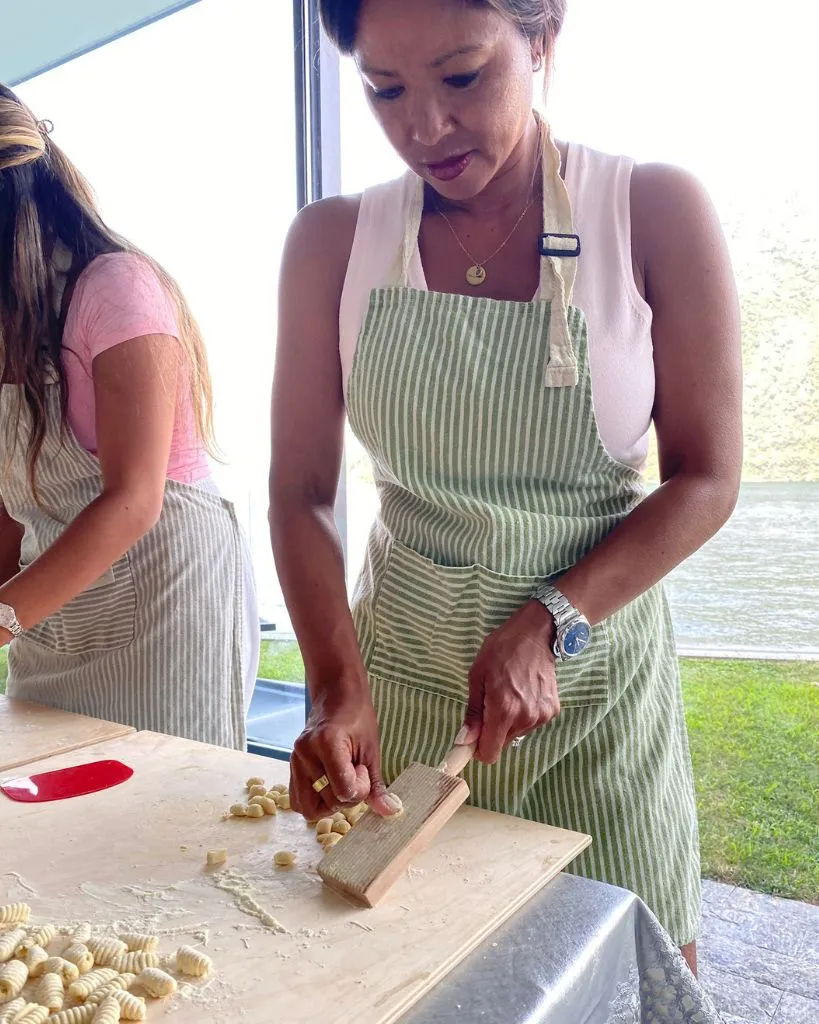 Two women in striped aprons preparing fresh gnocchi dough on a wooden board near a window.