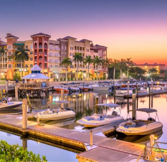 Marina at sunset with docked boats, palm trees, and colorful residential buildings.
