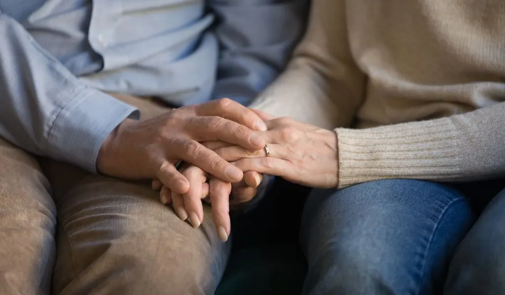 Close-up of two people sitting, one wearing a beige sweater and jeans, holding hands gently.