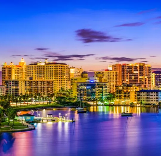 Nighttime cityscape with illuminated high-rise buildings reflecting on calm waterfront.