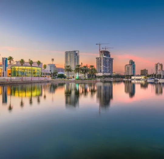 Calm waterfront cityscape at sunset with modern buildings and palm trees reflected in water.