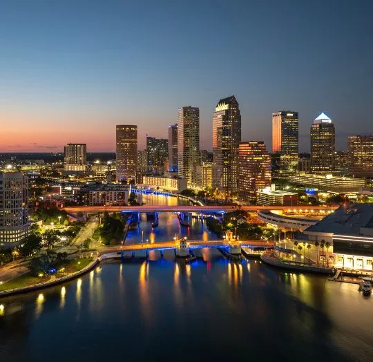 Aerial view of a city skyline at dusk with illuminated skyscrapers and bridges over a river.