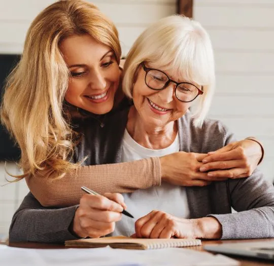 Middle-aged woman embracing elderly woman with glasses, both smiling and writing in a notebook.
