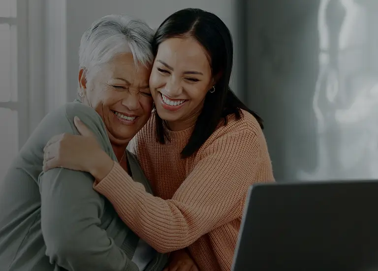 Two women, one older with gray hair and one younger with dark hair, smiling and hugging indoors near a laptop.