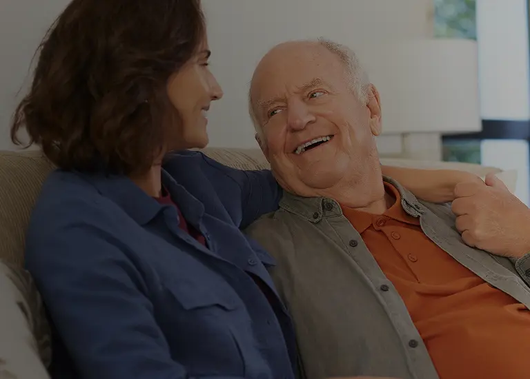 Elderly man and middle-aged woman smiling and sitting closely on a beige couch indoors.