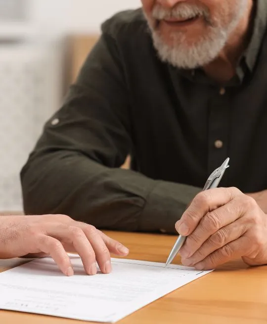 Two individuals, one elderly with a gray beard, reviewing and signing a document on a wooden table.