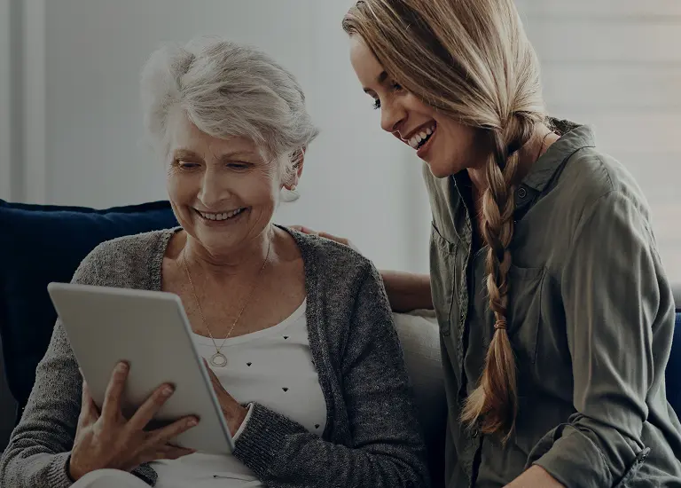Elderly woman and younger woman smiling while looking at a tablet together.