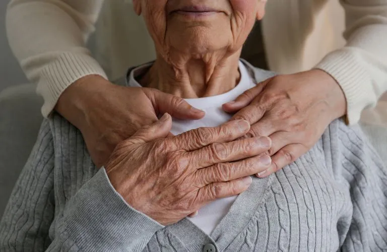 Close-up of elderly person wearing gray cardigan with hands of another person gently resting on their shoulders.