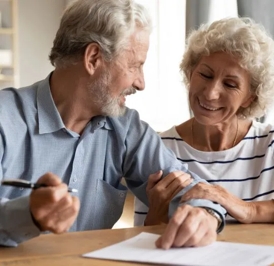 Elderly couple smiling at each other while reviewing a document at a wooden table.