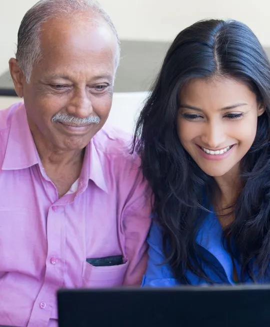 Two individuals, an elderly man in a pink shirt and a young woman in a blue top, smiling while looking at a laptop.