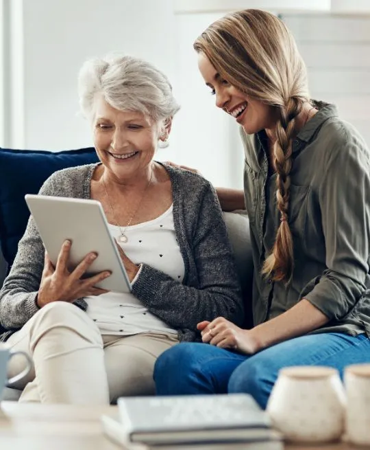 Two women, one elderly and one younger, sitting on a couch and smiling while looking at a tablet.