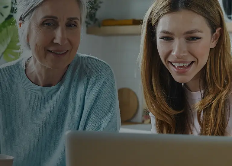 Two women, one older with gray hair in a light blue sweater and one younger with long brown hair, smiling while looking at a laptop.