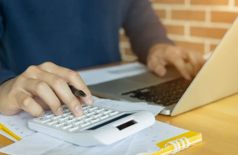Person using a white calculator and laptop on a desk with documents and a yellow notebook.