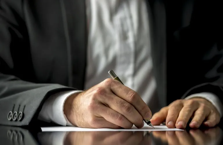 Close-up of a person in a suit writing on white paper with a pen on a reflective surface.