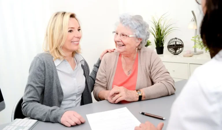 Two women, one elderly and one middle-aged, smiling and seated at a desk with a third person.