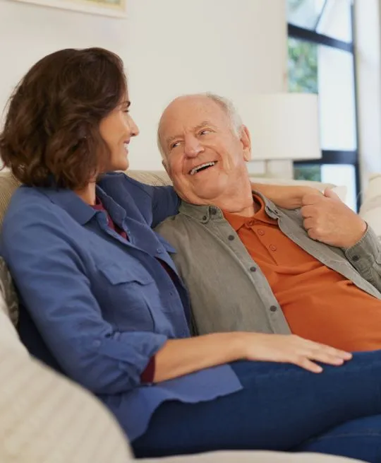 Middle-aged woman and elderly man sitting on a beige sofa, smiling and conversing indoors.