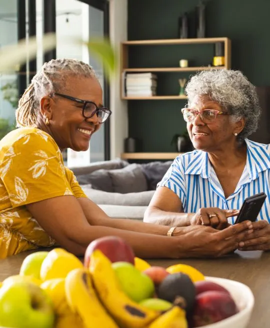 Two elderly women with glasses smiling and looking at a smartphone in a modern living room.