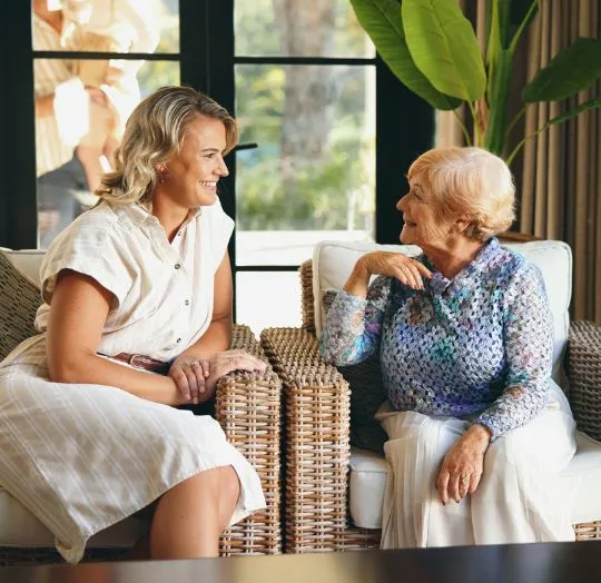 Two women, one elderly and one younger, engage in a friendly conversation on wicker chairs indoors.