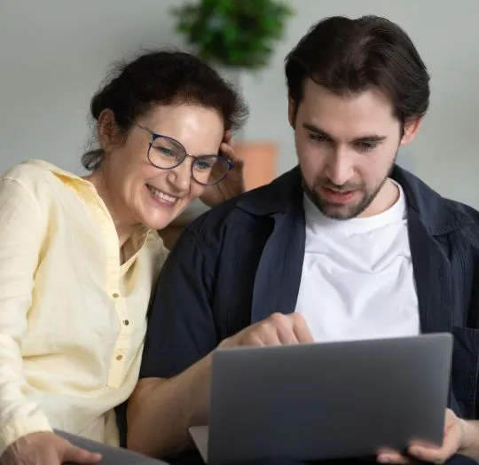 Two adults, a woman in a yellow shirt and a man in a white shirt and dark jacket, looking at a laptop screen together indoors.