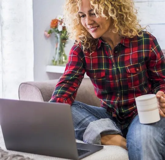 Woman with curly blonde hair in a red plaid shirt holding a white mug, using a laptop.